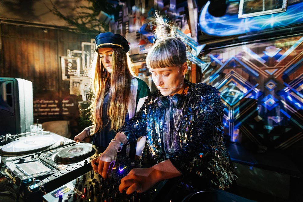 Two stylish DJs performing together late into the night at a colourful open air nightclub.