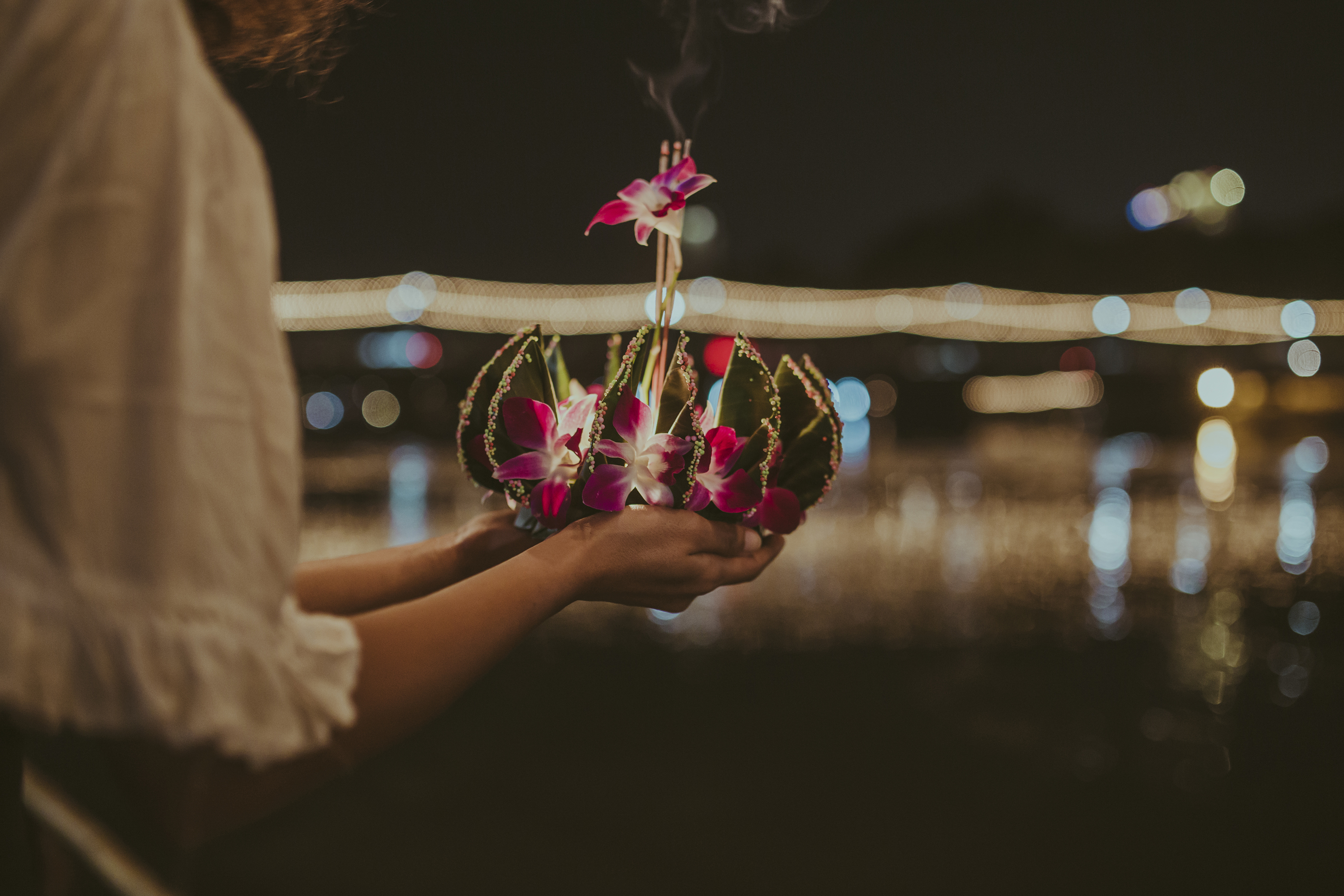 Person holding a decorated floating lantern with flowers and incense at night.