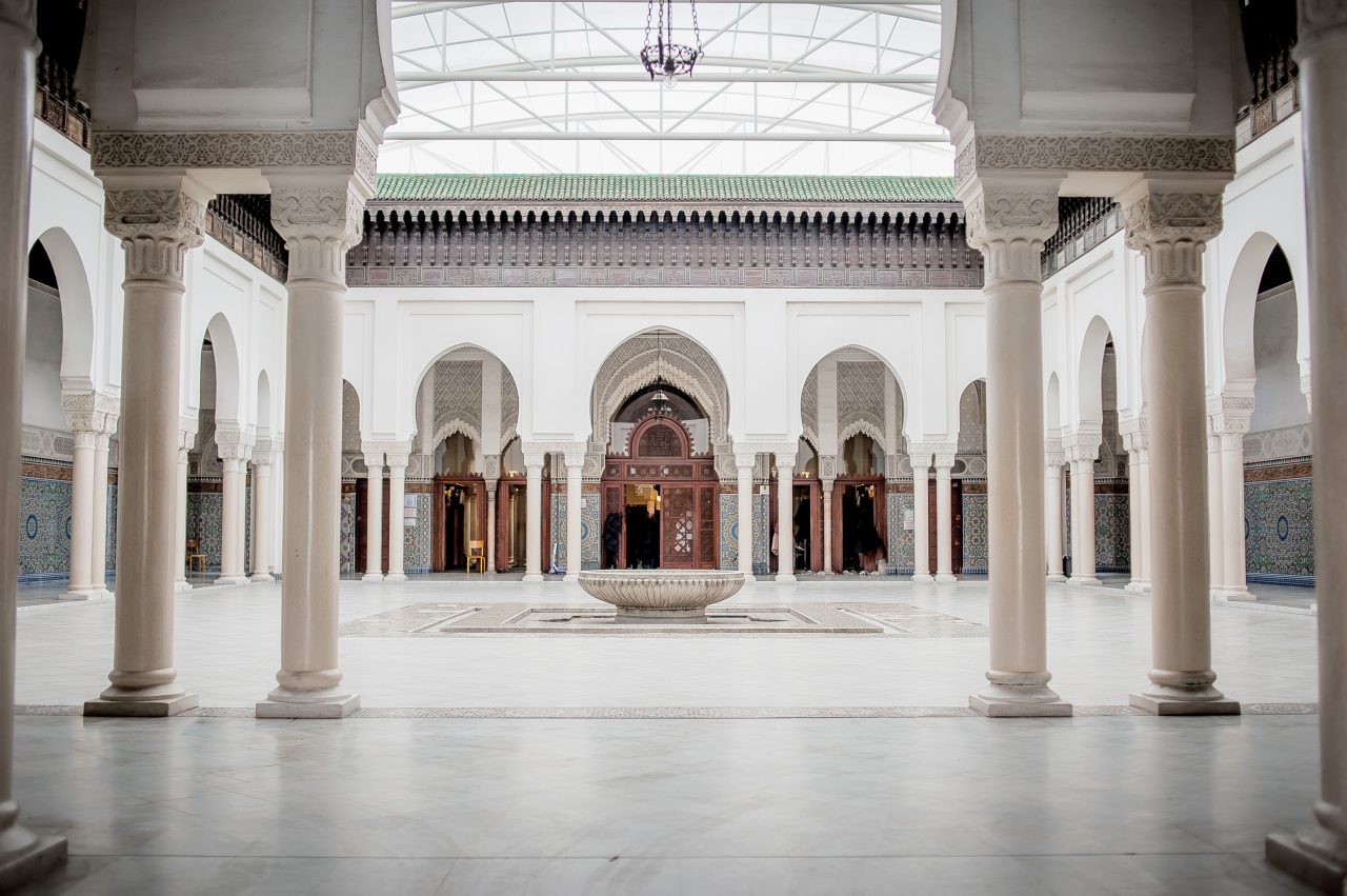 Interior courtyard of a grand Moorish-style building with ornate arches, carved columns, patterned tile walls, and a central stone fountain beneath a glass-covered roof.