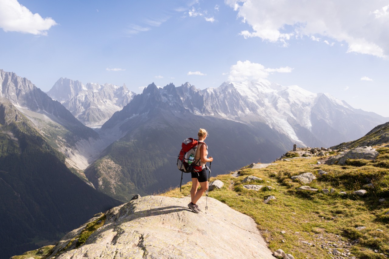 A young woman is hiking with trekking poles in the French Alps mountains overlooking Mont Blanc peak.