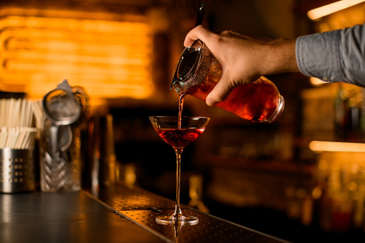 wine glass standing on bar in which male hand pours a drink from a mixing cup with a strainer