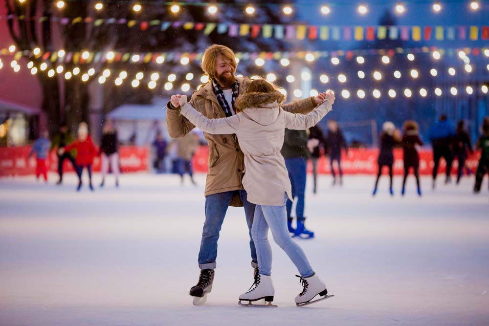 Theme ice skating rink and loving couple. meeting young, stylish people ride by hand in crowd on city skating rink lit by light bulbs and lights. Ice skating in winter for Christmas on ice arena.