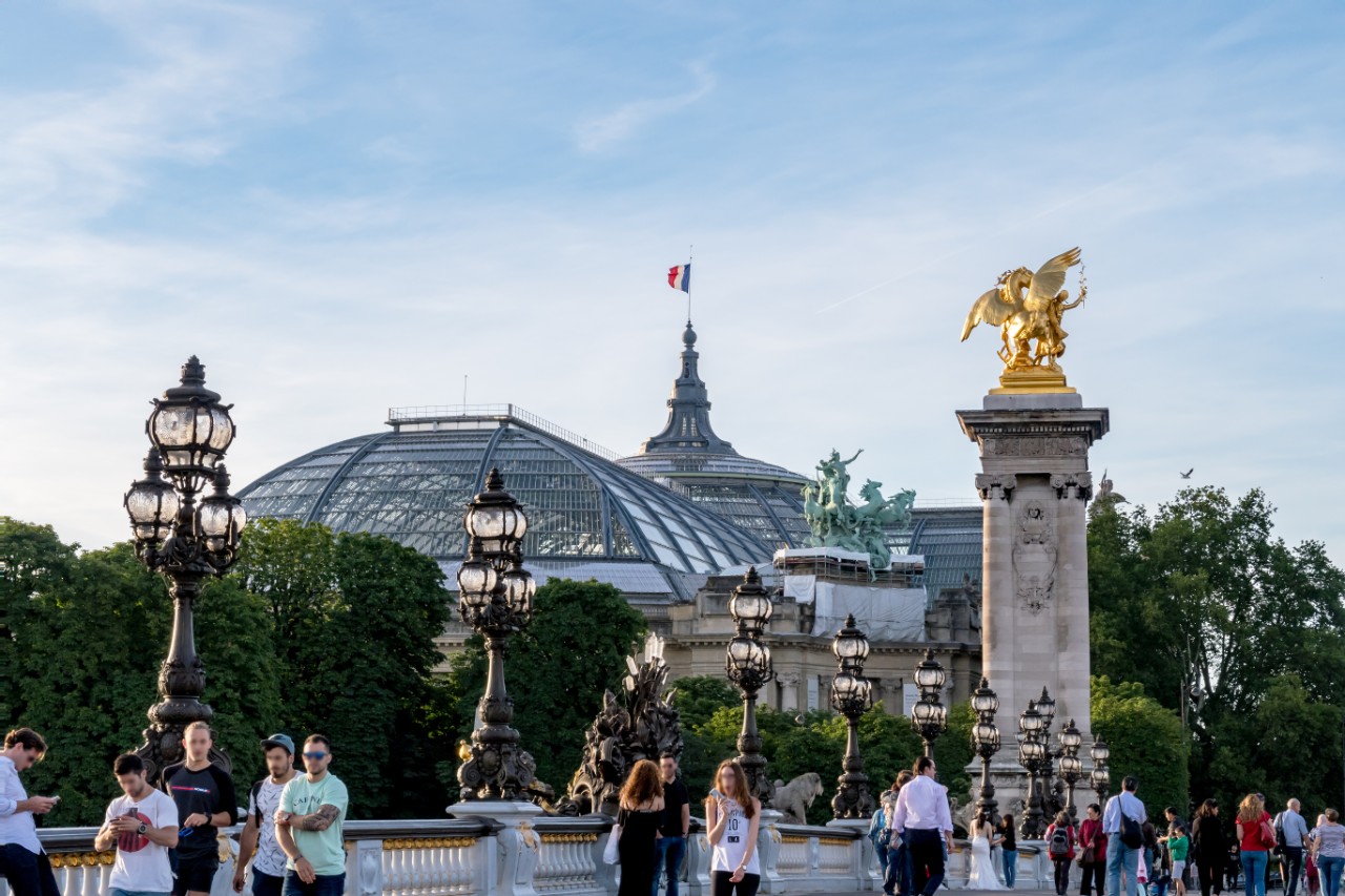 Pedestrians wandering on Pont Alexandre III on Seine River with Grand Palais in background.